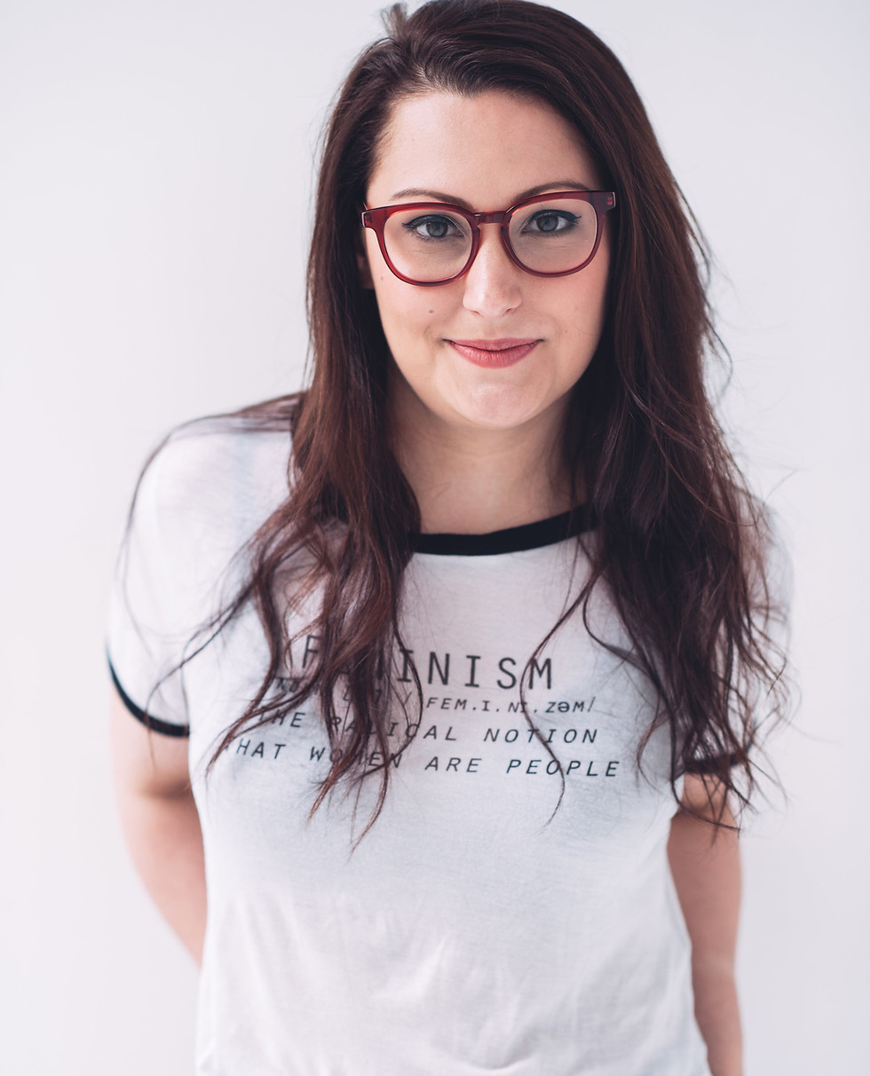 Delaney Amatrudo, MFT is pictured wearing red eyeglasses and a t-shirt that reads, "Feminism: The radical notion that women are people."