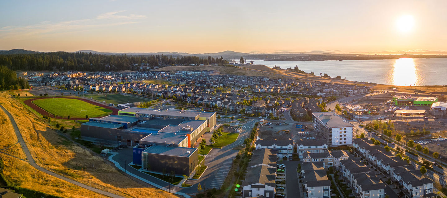 Royal Bay master-planned community in Colwood, BC — aerial view at sunset showing Royal Bay Secondary School, residential neighbourhoods, and the Strait of Juan de Fuca shoreline