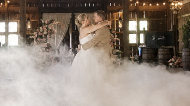 A bride and groom share their first dance in a dimly lit reception hall while thick, white dry-ice clouds swirl around their feet, creating a dreamy ‘dancing on a cloud’ effect. Guests watch from the perimeter as soft purple lighting adds to the romantic atmosphere.
