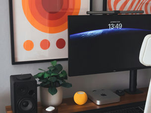 A home office desk setup with a minimalist and artistic aesthetic. On the left, a framed "BAUHAUS" exhibition poster with concentric orange and red circles hangs on a light gray wall. To its right, another framed print with wavy orange and white lines is visible. The wooden desk holds a black computer monitor displaying a blue abstract background and the time "13:49". A monitor light bar is attached to the top of the screen. To the left of the monitor, there's a black speaker and a small green plant in a white pot. A yellow textured ball and a silver mini-computer are also on the desk. On the front left of the desk, there's an orange-red book titled "JEMNE UMENIE NENECHAT SA SRA*T" and a small white object that looks like a candle or diffuser. A white ergonomic office chair is pulled up to the desk on the right side of the image. The photo is by Matúš Gocman on Unsplash.