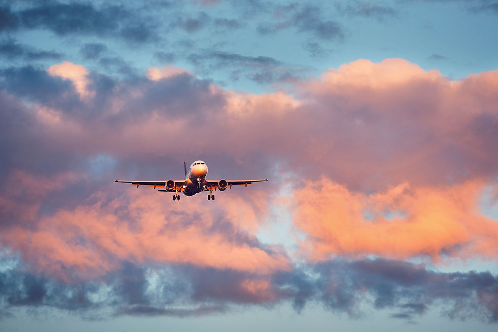 A head-on shot of a commercial passenger airplane descending for a landing, seen from directly below its flight path. The aircraft has its landing gear extended and its headlights illuminated against a dramatic sunset sky filled with dense clouds painted in shades of pink, orange, and deep slate blue.