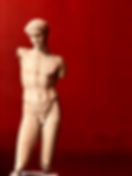 A stunning classical age statue on display at the National Archaeological Museum of Athens, set against a deep red wall. The statue depicts a human figure in a pose of grace and strength, with intricate details and lifelike proportions. The warm lighting highlights the texture and color of the statue, while the bold red background creates a sense of drama and contrast.