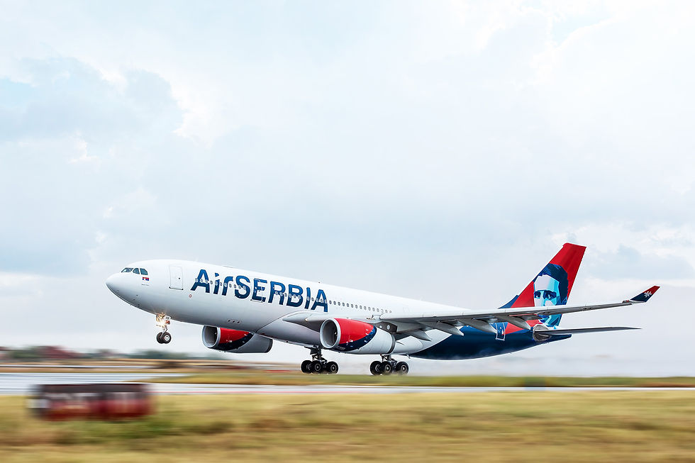 An Air Serbia Airbus A330 commercial passenger airplane is captured mid-takeoff, lifting off from an airport runway. The wide-body aircraft features a white fuselage with large blue "AirSERBIA" lettering, red engine nacelles, and a distinctive red and blue tail fin featuring a stylized portrait illustration. The plane is angled upward against a bright, cloudy sky, with a motion-blurred grassy field and runway markings visible in the lower foreground.
