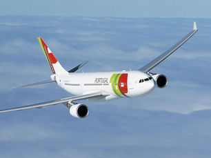 An aerial, eye-level shot of a white TAP Air Portugal commercial passenger airplane banking slightly to its left while cruising high above a thick layer of soft, white clouds under a clear blue sky.