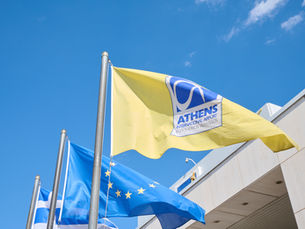 A yellow Athens International Airport flag waving on a flagpole alongside the European Union flag outside a modern terminal building under a clear blue sky.