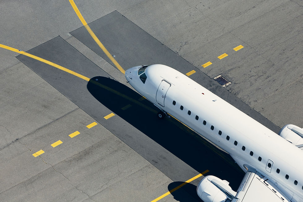 A high-angle, top-down view of a modern white narrow-body commercial airplane idling on an asphalt airport tarmac. The nose and forward fuselage of the aircraft cast a sharp, dark shadow to the left over painted yellow taxiway lines and markings. Part of the plane's right engine and wing are visible at the bottom of the frame under bright daylight.
