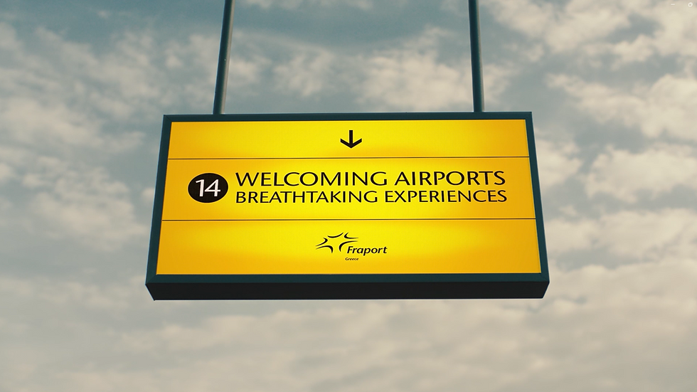 Here is the alt text for the image:
A rectangular, yellow airport-style sign hanging against a cloudy sky backdrop. The sign features a downward-pointing arrow at the top. The middle section contains a black circle with the number "14" inside, followed by the text "WELCOMING AIRPORTS BREATHTAKING EXPERIENCES" in all-caps, black lettering. The bottom section displays the "Fraport Greece" logo.