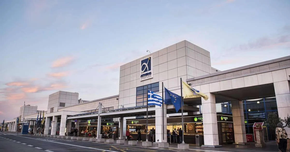 A wide-angle view of the bright and airy main terminal at Athens International Airport, featuring sleek lines, soaring ceilings, and plenty of natural light. Crowds of travelers can be seen moving through the space, with check-in desks, security scanners, and shops and restaurants visible in the background.