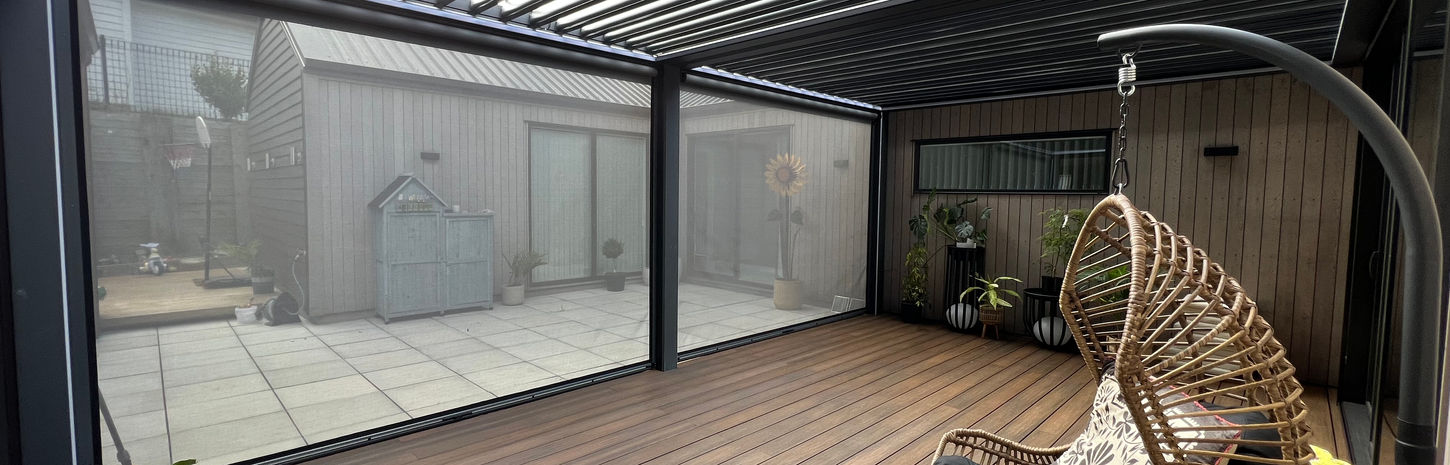 An open Skyline Louvre and closed cream outdoor blinds let the sunlight into a wood deck with an egg chair in the foreground. 