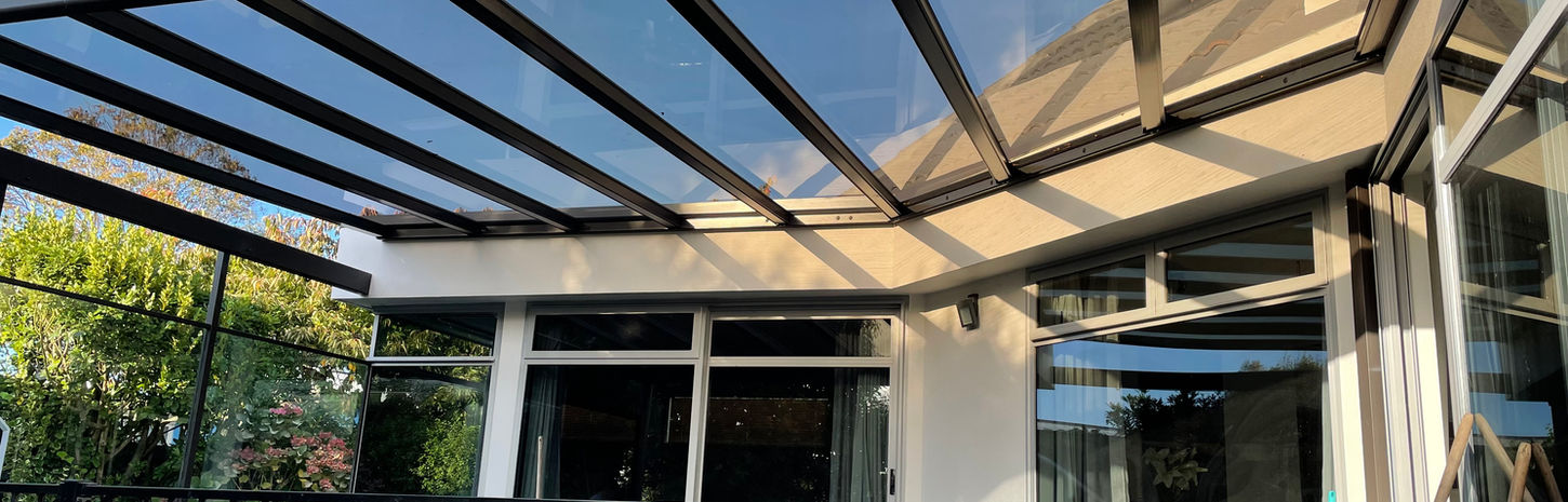 A clear Transroof with black framing enclosed on 3 sides by the curved roof of a house. The patio underneath it is enclosed by a fence. 