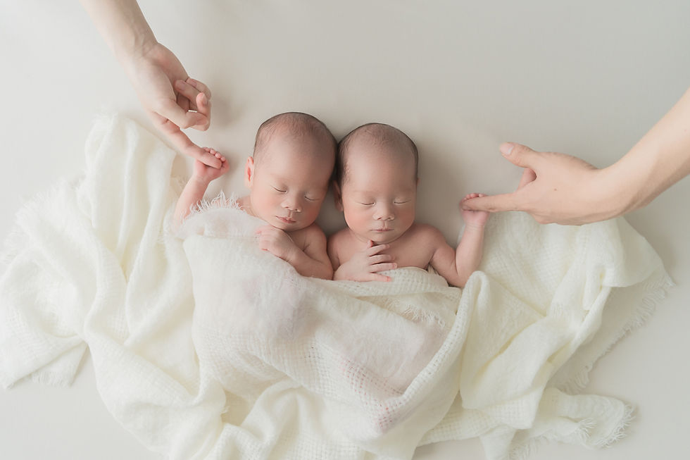 白い布に包まれ眠る双子のニューボーンフォト。大人の手が添えられている。