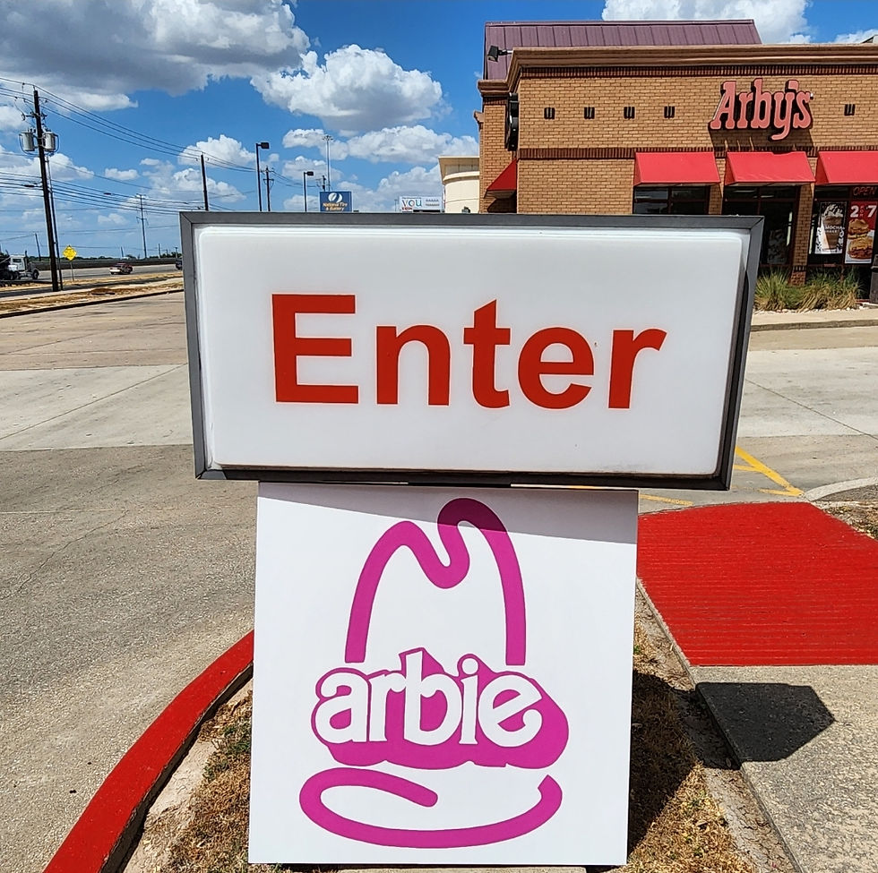 ARBY #2 (2023) displayed in front of a beautiful Arby's restaurant. The painting, which says "arbie" in pink letters, is propped in front of a sign that says Enter. 