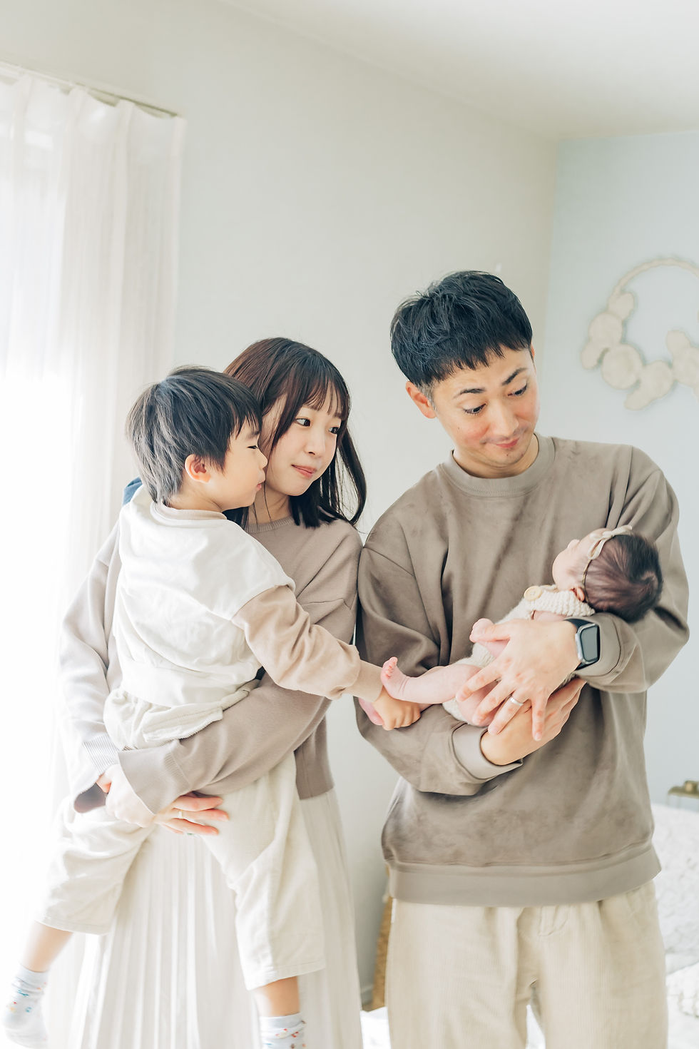 若い夫婦と幼児、乳児がリビングにいる。母親は幼児を抱き、父親は乳児を見つめて微笑んでいる。明るい雰囲気。