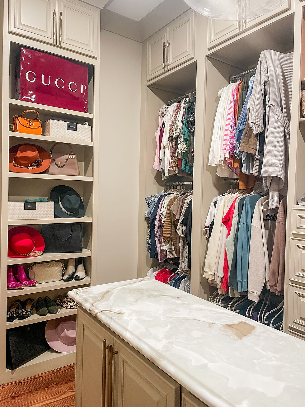 Walk-in closet with clothes on racks, shelves of hats and shoes, a marble-topped island, and a visible large red Gucci bag. Cozy and organized.