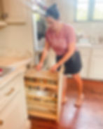 "SOUTHERN SORT PROFESSIONAL ORGANIZING" is organizing a spice rack in a kitchen. The spice rack is a pull-out drawer filled with many small spice jars. Below the spice jars are shelves with bottles. The kitchen has light-colored cabinets, a light-colored countertop, and a wooden floor.