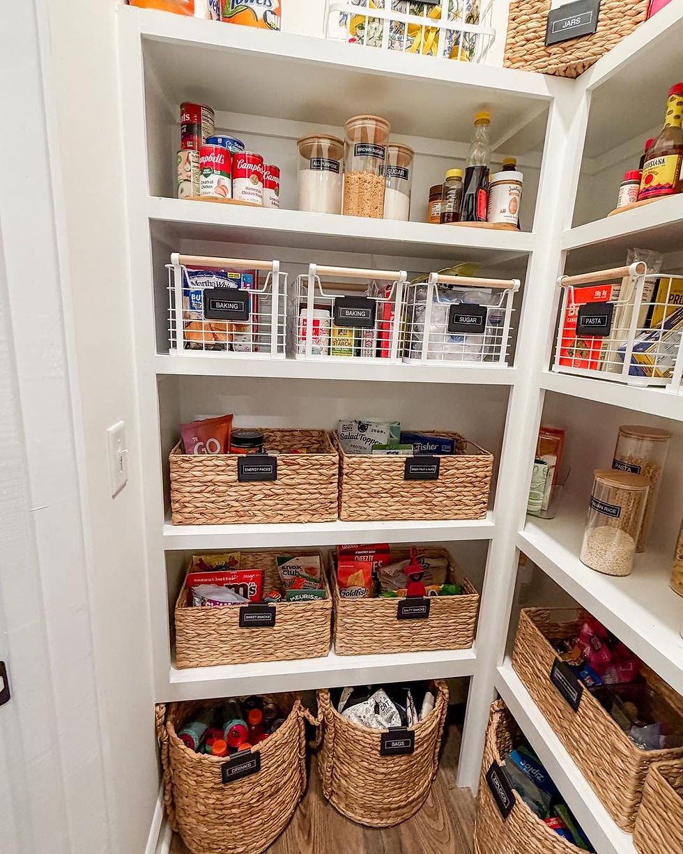 Neatly organized pantry with labeled baskets and jars of baking goods, snacks, pasta, and drinks on white shelves.