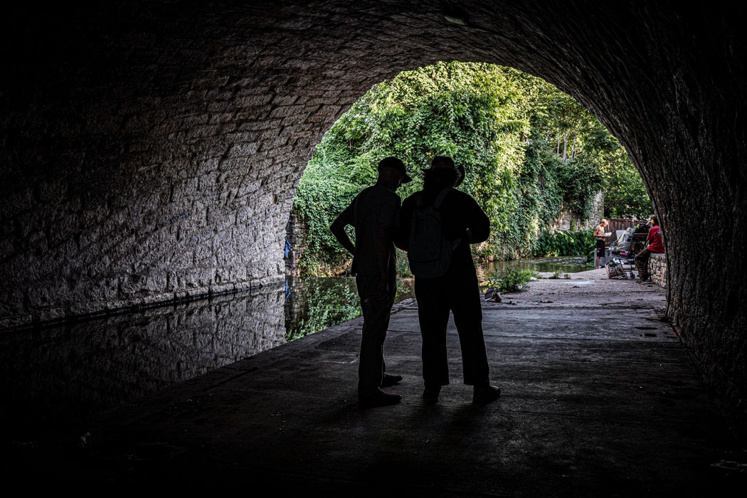 Two figures in silhouette inside a stone tunnel that opens onto a green creek bed.