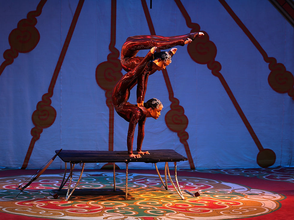 Dos acróbatas en traje rojo brillante hacen una figura contorsionista sobre una mesa, en un escenario azul con luces cálidas.