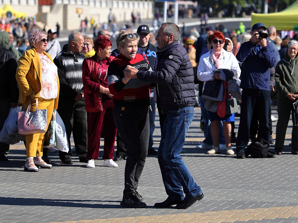 Pareja mayor baila en una plaza al aire libre, rodeada de gente; ella viste rojo y negro, él chaqueta azul; ambiente festivo.