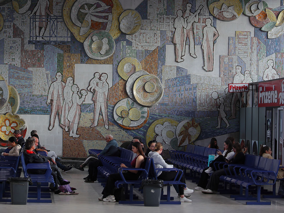 Personas sentadas en bancos azules frente a un gran mural de mosaico; se ve el letrero Frizerie y un ambiente de espera.