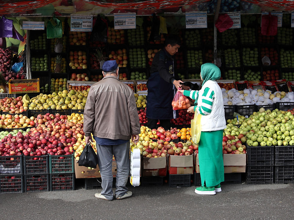 Mercado de frutas al aire libre: vendedor entrega una bolsa a una clienta; otro cliente espera entre montones de manzanas y carteles.