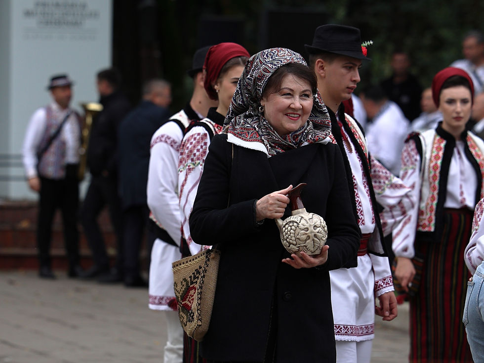 Mujer sonriente con pañuelo y abrigo negro sostiene una vasija; detrás, grupo con trajes tradicionales blancos y rojos.