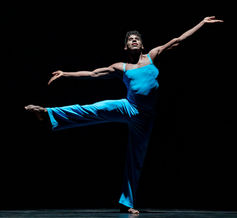 A solo dancer in a bright blue costume balances gracefully on one leg, arms extended wide, illuminated against a dark stage, showcasing strength, poise, and control.