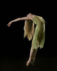 A lone dancer in a flowing green dress arches backward on pointe, hair cascading, body suspended in a luminous curve against the black void.