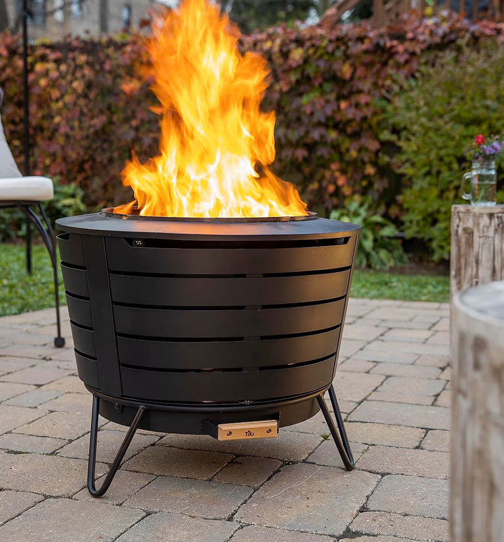 Tall orange flames rise from a black Tiki fire pit on a stone patio, with green shrubs and a chair in the background.