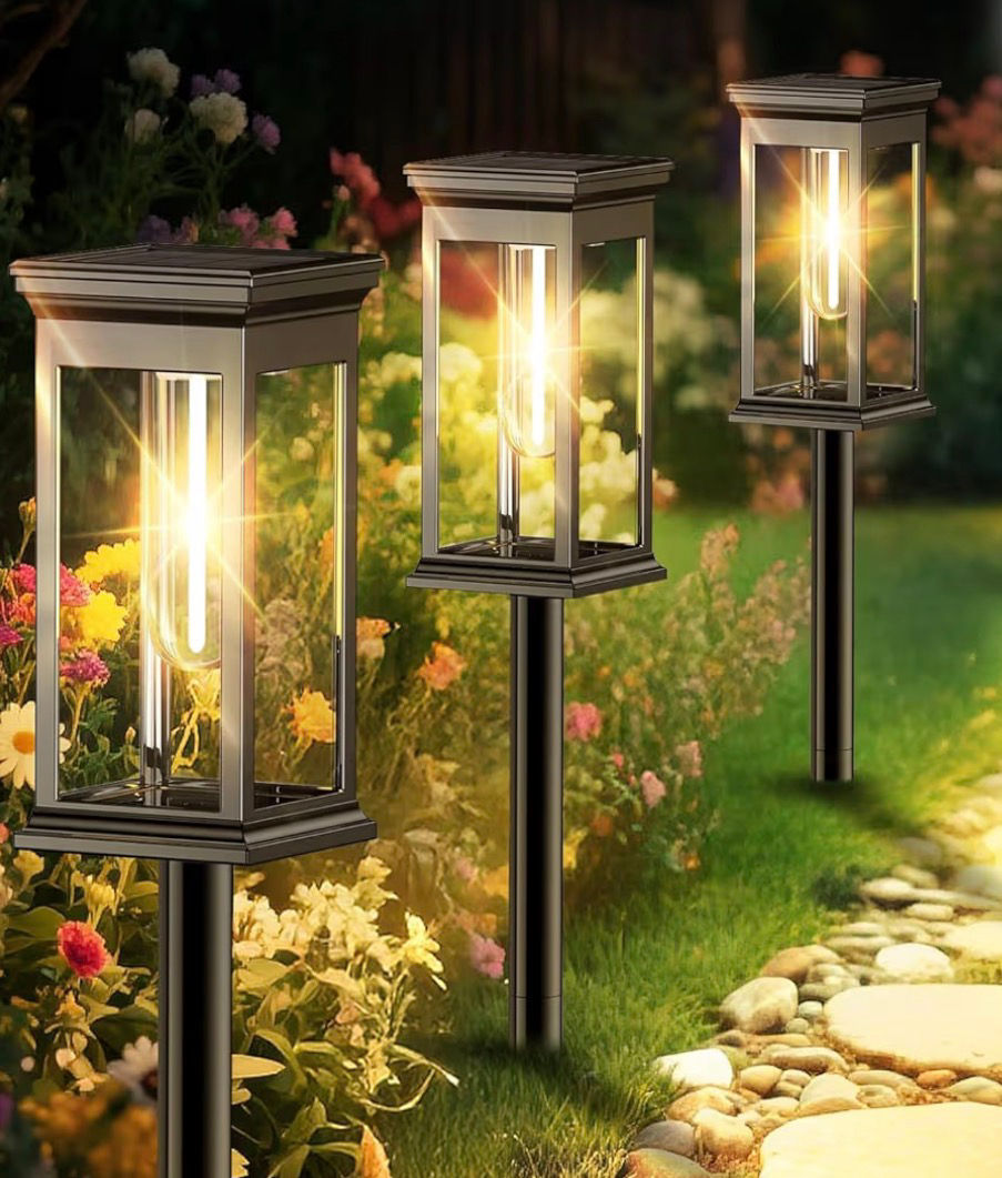 Three glowing black lantern-style garden lights line a flower-filled path at dusk, with warm golden light and a stone walkway.