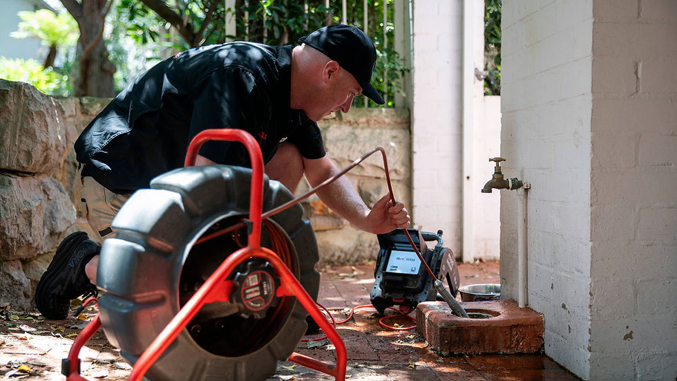 Mark Lawler is using a drain CCTV to check outdoor plumbing. Surrounded by bricks and foliage. Focused atmosphere amid green and brown tones.