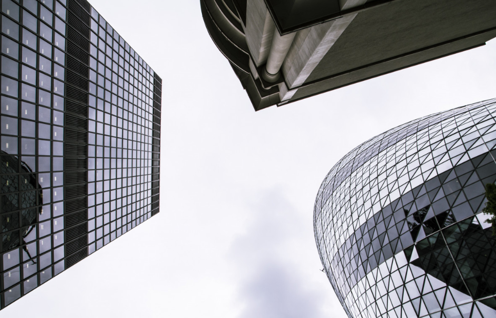 Looking up at modern skyscrapers with glass facades in a cloudy sky, showing geometric patterns and urban architecture.