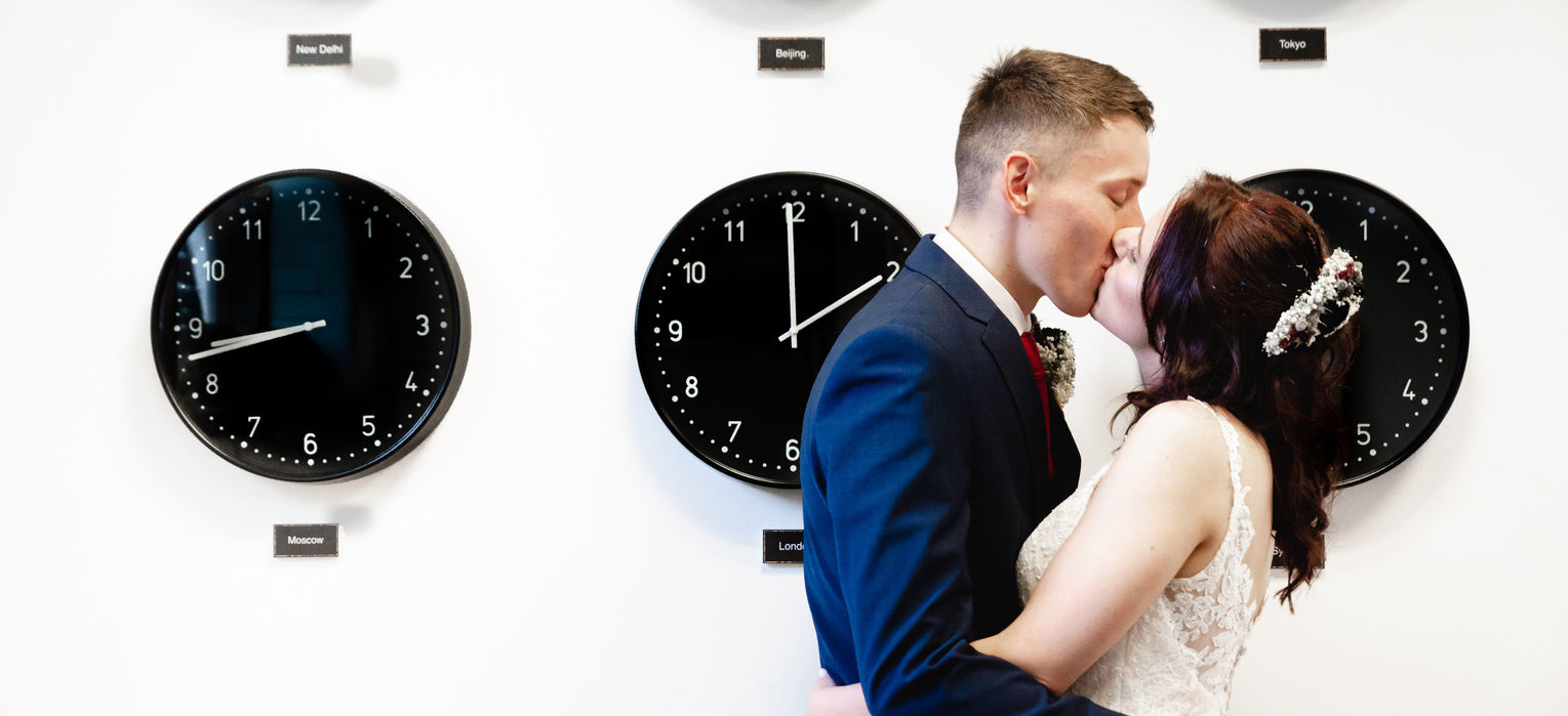 A couple kissing, the background is white wall with clocks showing time from all over the world.