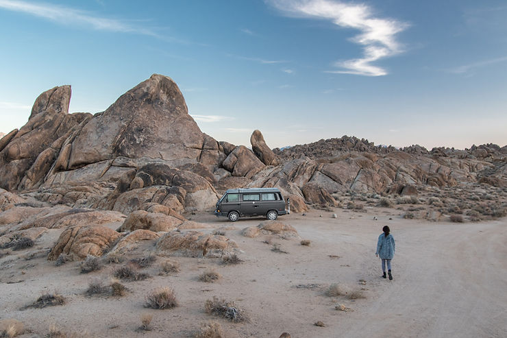 An old camper van parked in a rocky terrain. A man walking towards his van.