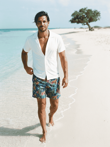 Model walking on the beach in Aruba wearing Sun Kulture Eco Swim Short in color Palm Island Fiesta and Blanco Breeze Cabana Shirt