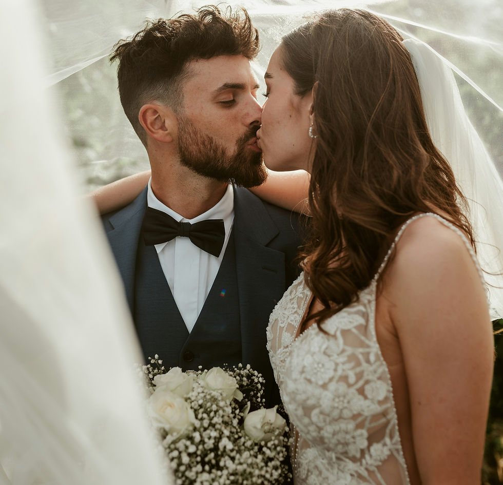 A newlywed couple shares a tender kiss under a veil, the bride holding a bouquet of white roses, capturing a moment of love and happiness on their special day.