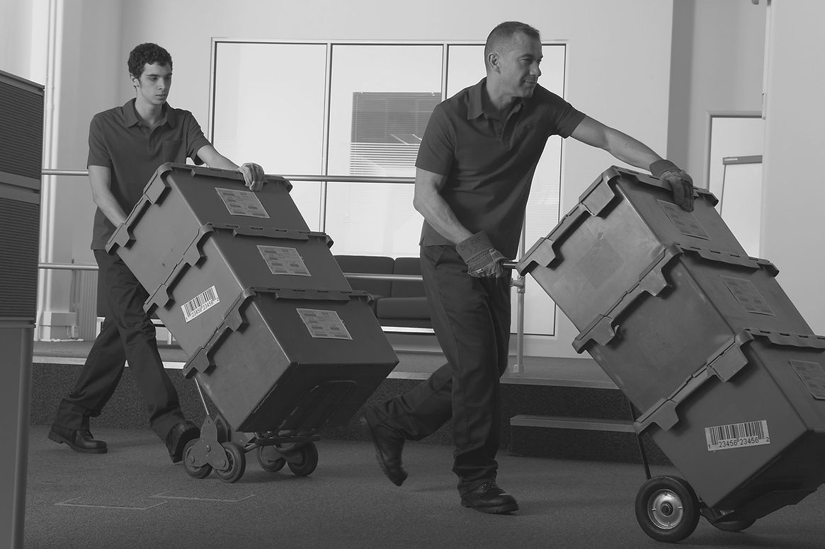 Two men wheeling crates on sack barrows through office area