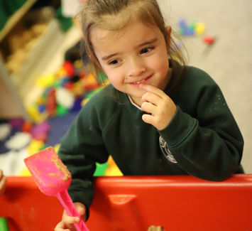 Girl playing in sandpit with pink shovel, looking thoughtful.