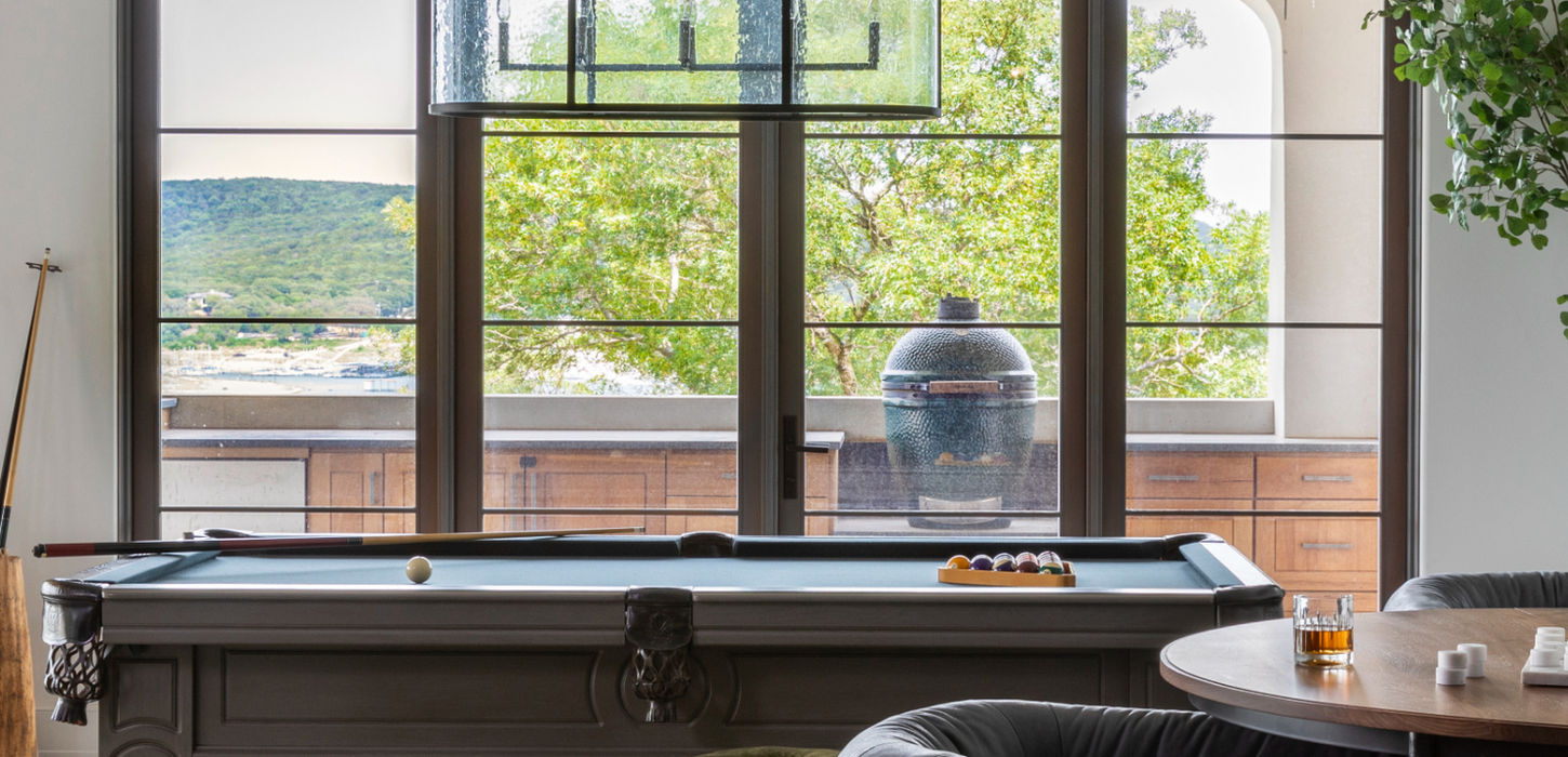 A stylish game room with a pool table, plush seating, natural stone flooring, and large windows showcasing expansive outdoor views. The space balances leisure, comfort, and understated elegance. Located in Austin, Texas. Interior design by Blair Burton Interiors.
