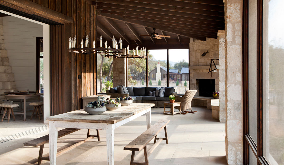 A spacious indoor-outdoor living area with a rustic dining table, wooden ceiling, stone fireplace, and comfortable lounge seating that encourages gathering and slow living. Natural materials blend seamlessly with surrounding views. Located in Austin, Texas. Interior design by Blair Burton Interiors.