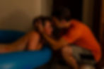 Man in orange shirt gently supports a woman in a blue inflatable birthing pool. The setting is dimly lit, conveying a calm, intimate mood.