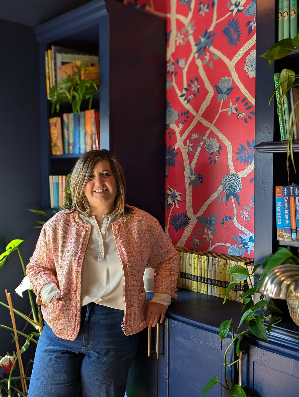 Lillian Medhus smiling, wearing a pink jacket in a room with dark blue shelves and floral wallpaper. Books and plants decorate the background.