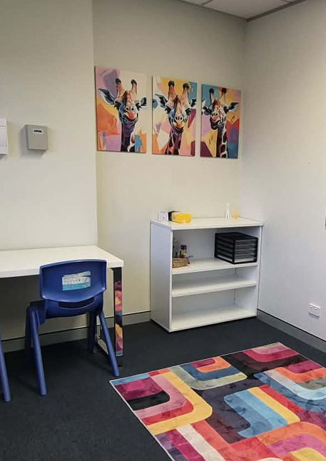 A therapy room with a colourful abstract rug, whiteboard, desk with two blue chairs and three abstract artworks featuring giraffes on the wall.