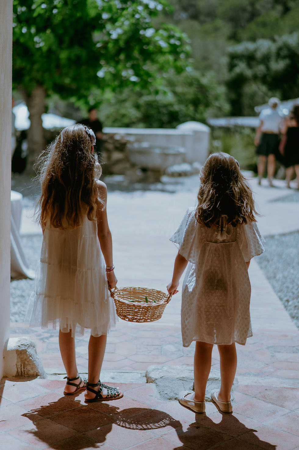 Flower girls in white cotton dresses holding a basket between them, photographed from behind.
