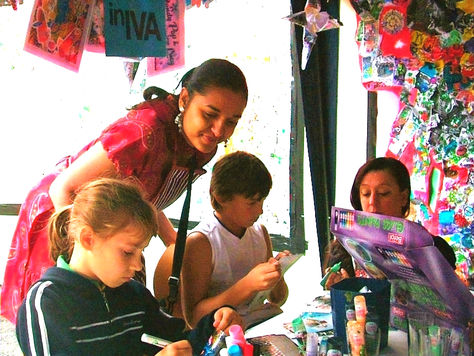 A group of three children, including a young girl in a dark blue jacket, are creating artwork with markers and glitter glue at a table, while a south Asian woman artist watches - engaged.