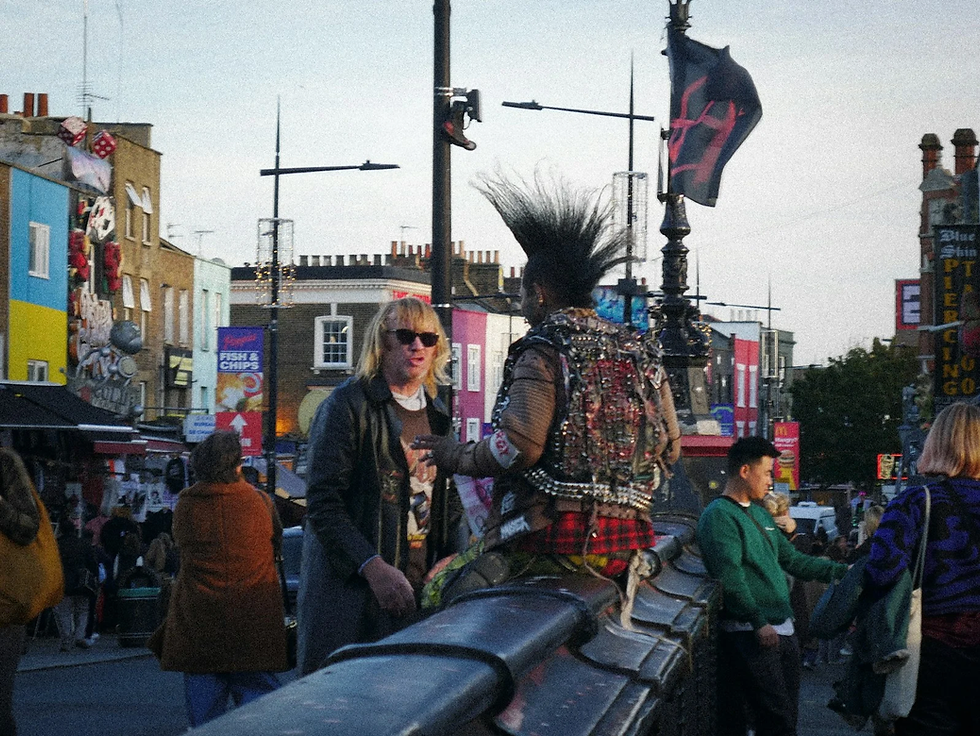 Punk man with mohawk and studded jacket chats with a blond man in sunglasses on a colorful street lined with signs.