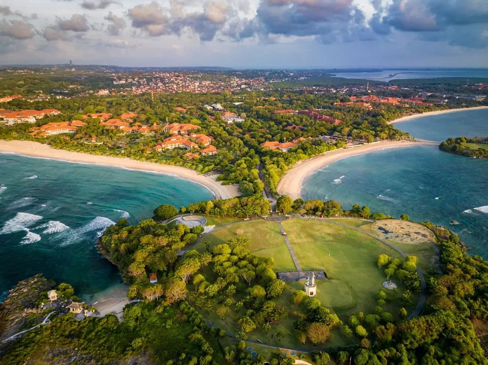 Aerial view of Nusa Dua, Bali showcasing twin curved beaches, lush greenery, luxury resorts with terracotta roofs, and the calm turquoise ocean under a dramatic sky.
