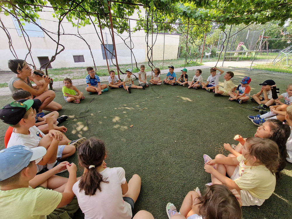 Copii și un adult stând în cerc la o activitate în aer liber.
