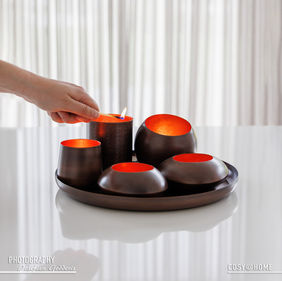 Sfeerfoto van een set theelichthouders voor kaarsen op een weerspiegelend tafelblad voor een doorzichtig gordijn. Een hand steekt een kaars aan met een lucifer.