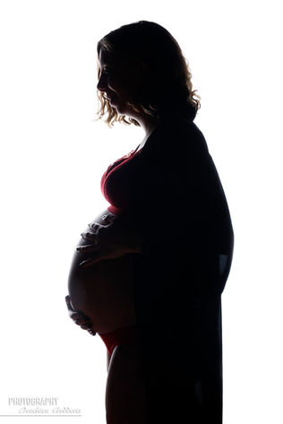 Silhouet van een zwangere vrouw in lingerie, met haar handen beschermend om haar buik gevouwen, tegen een fel verlichte achtergrond.